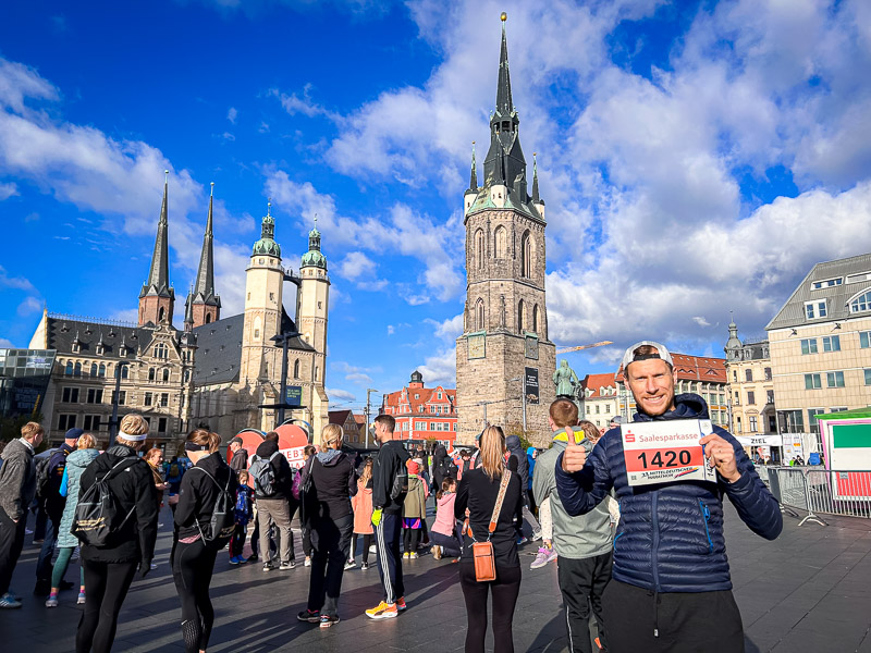 A participant of the Mitteldeutscher Halbmarathon holds up his bib number on Halle's (Saale) marketplace. Behind him, the historic Roter Turm and Marktkirche are visible. In the foreground, runners and spectators prepare for the race.