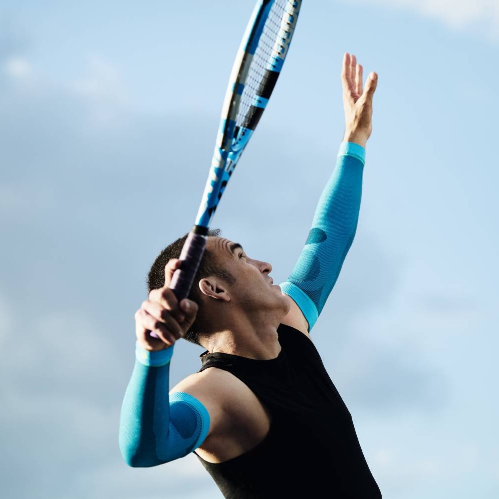 Tennis player with blue compression sleeves serving A tennis player wearing blue compression sleeves serving. The sky is blue with a few clouds.
