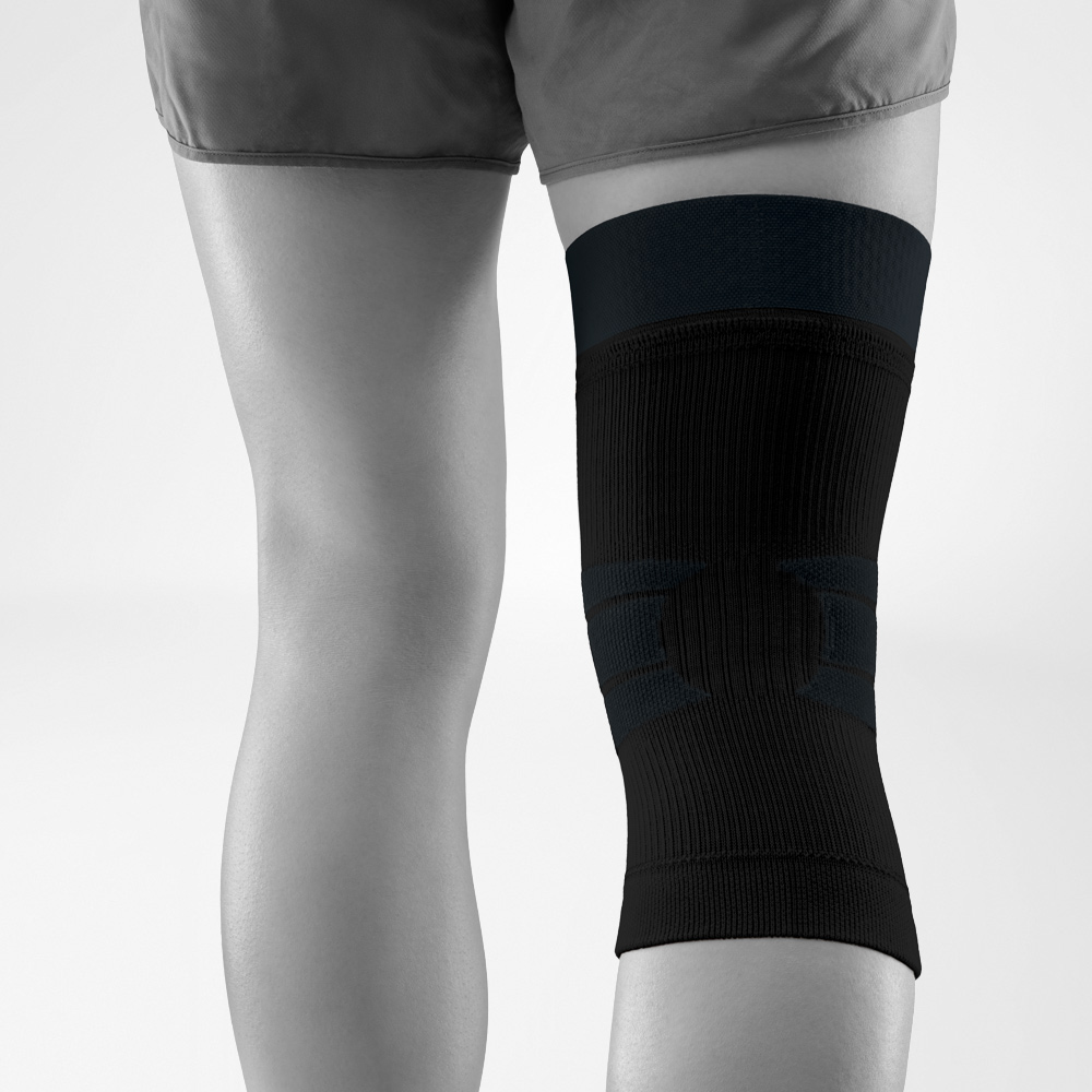 Back view of a worn black knee support. The anatomical knit design with integrated stabilization surrounds the knee securely while allowing freedom of movement outdoors.