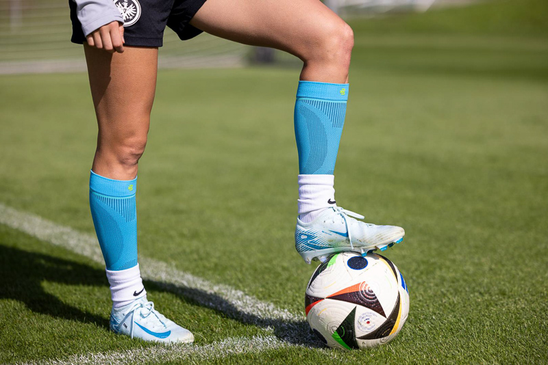 Close-up of a football player wearing Bauerfeind compression socks, resting one foot on a ball during practice. The green pitch and crisp details highlight athletic performance and high-quality gear.
