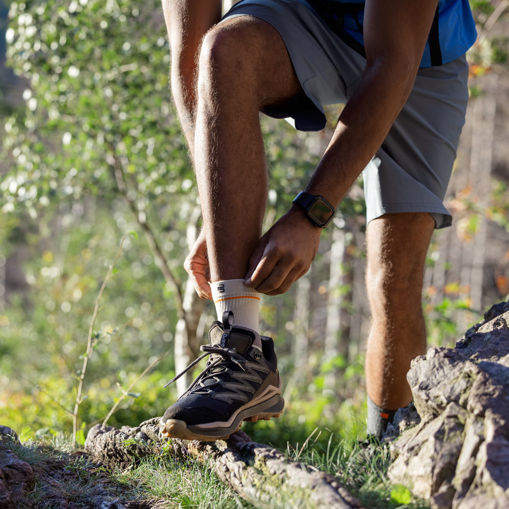 hiker stands on a narrow forest trail while adjusting the Outdoor Compression Ankle Support around his ankle. He is wearing sturdy hiking shoes, shorts, and a backpack.