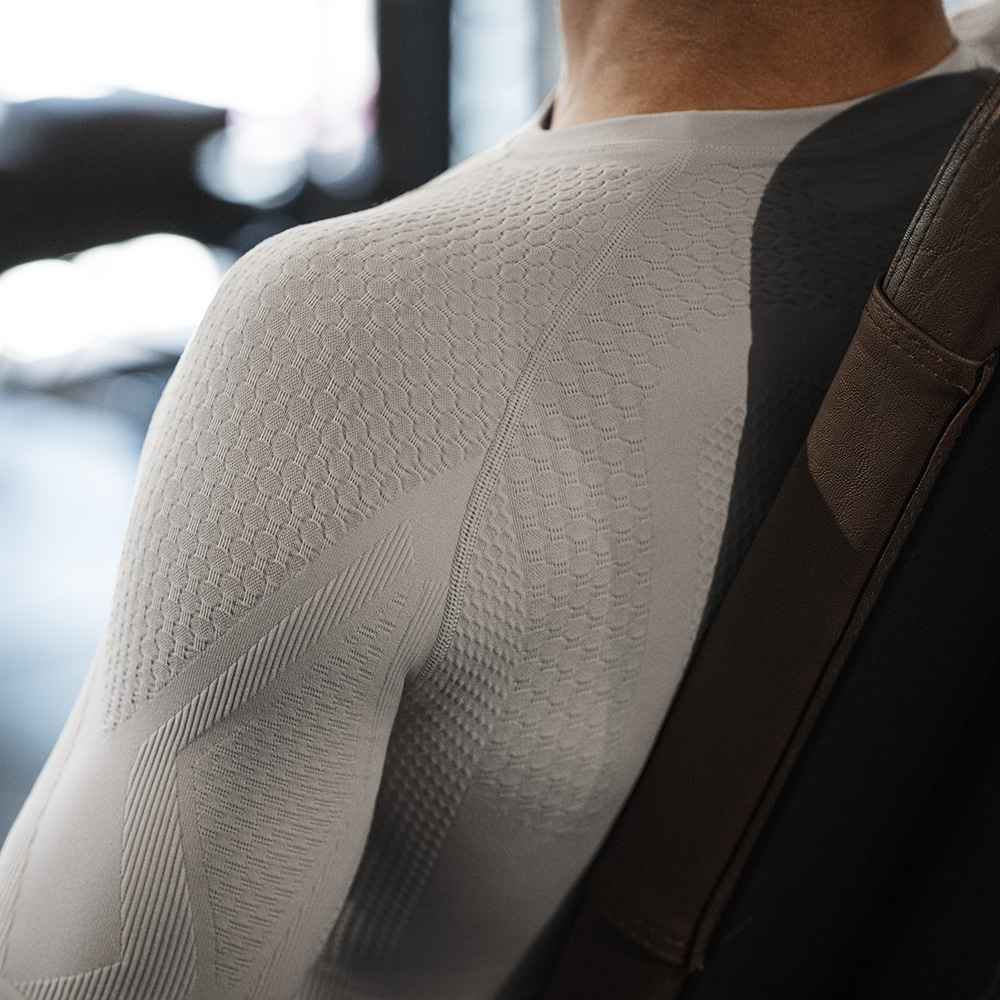 Close-up of a man's right shoulder in a tight-fitting beige compression shirt with functional zones for targeted muscle support and maximum freedom of movement, captured in a sporty environment.