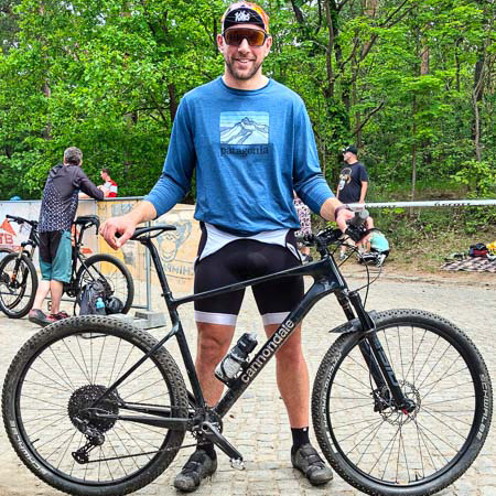 MTB-Grinsen: Nach der Challenge ist vor der Challenge! Ein lächelnder Mann steht mit einem schwarzen Cannondale-Gravelbike auf einem Waldweg. Er trägt eine blaue Patagonia-Langarmshirt und eine schwarze Radlerhose. Im Hintergrund sind andere Radfahrer und grüne Bäume zu sehen.