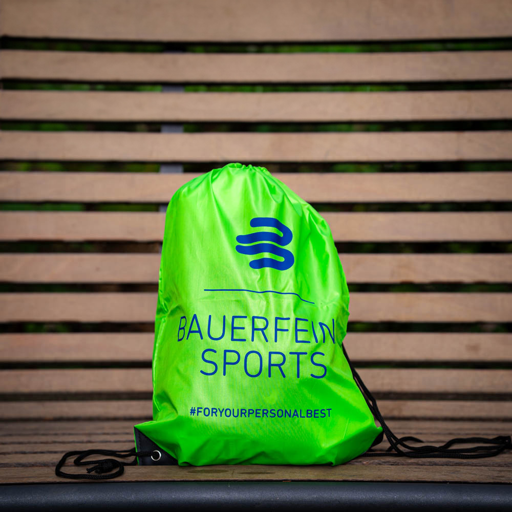 A green Bauerfeind Sports drawstring bag sits on a wooden bench, displaying the logo and hashtag #foryourpersonalbest.