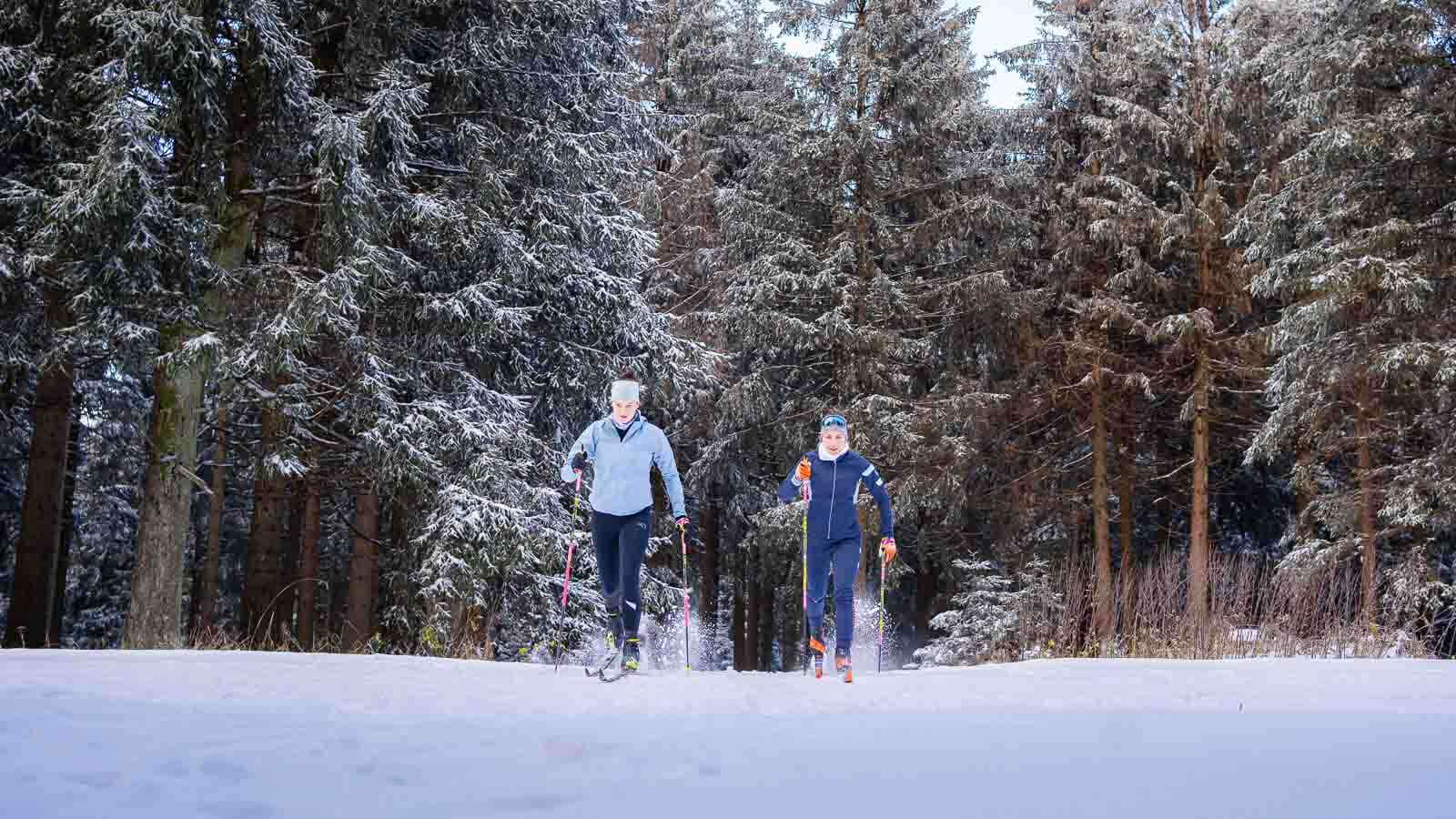 Two people cross-country skiing through a snowy landscape. They are wearing warm functional clothing, winter hats, and using ski poles. The surroundings are characterized by snow-covered trees and a crisp winter atmosphere.