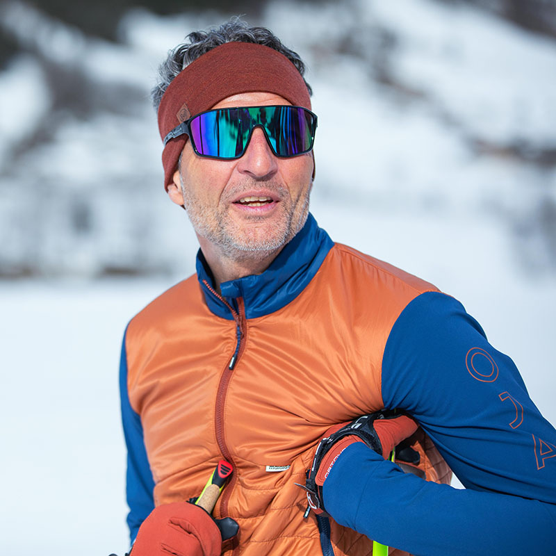 An elderly man wearing ski goggles, a brown headband and an orange-blue winter jacket was photographed in a portrait in front of a white forest.