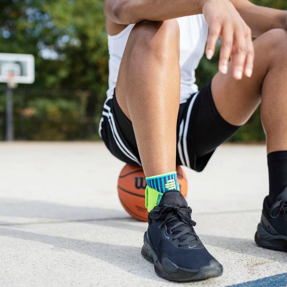 Stable on the court – with secure support! A basketball player sits on the court with a basketball under his leg. He is wearing the Bauerfeind Sports Ankle Support Rivera in blue-green on his ankle.