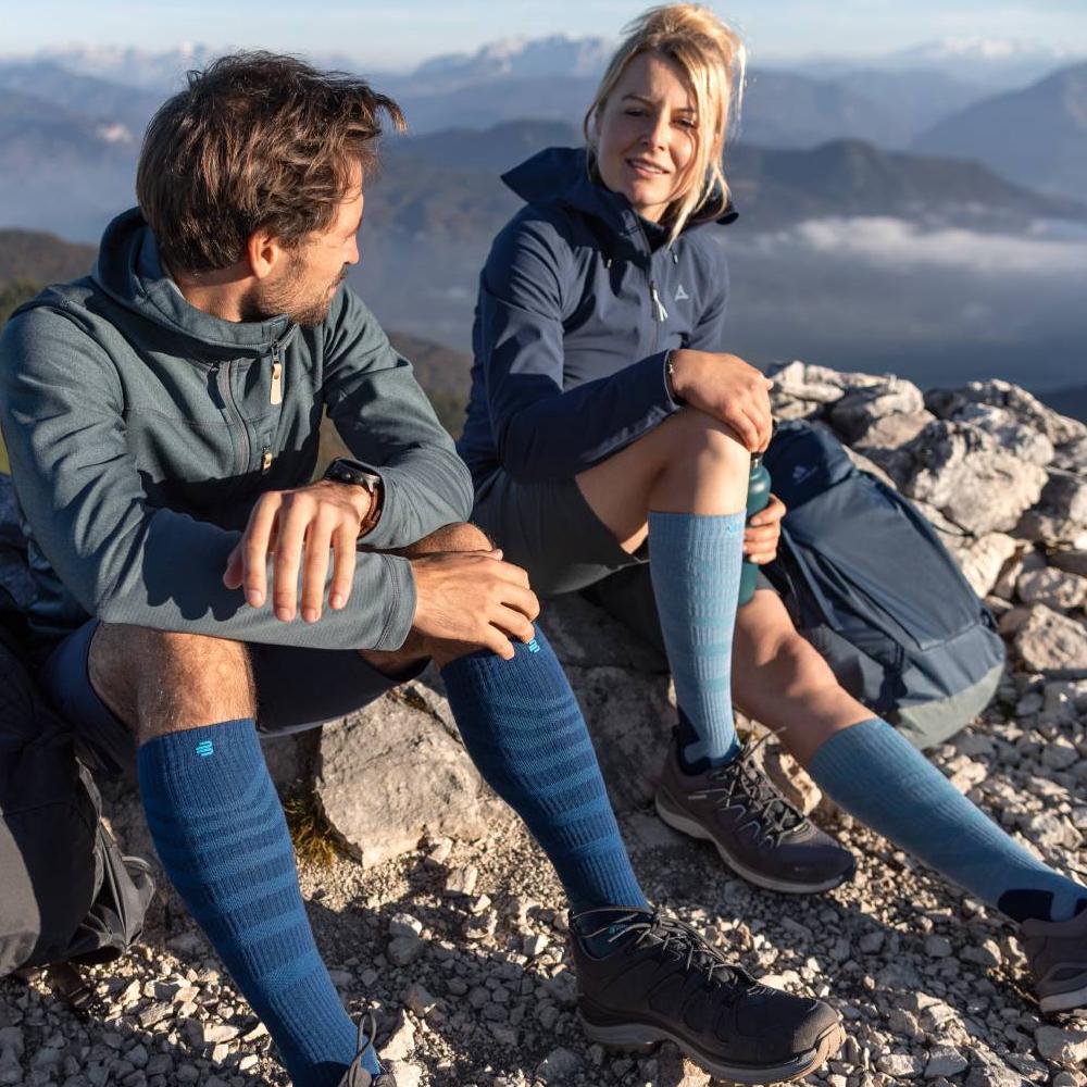 Reach new heights – comfort on every summit. Two hikers enjoying the view on a mountain summit while wearing Merino Outdoor Compression Socks in red-brown. The socks provide optimal comfort and support for long treks in challenging terrain.