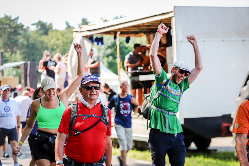 Happy participants celebrate during the 4Daagse walk in front of a music stage. A woman in a green top and a man in a red shirt walk by smiling.