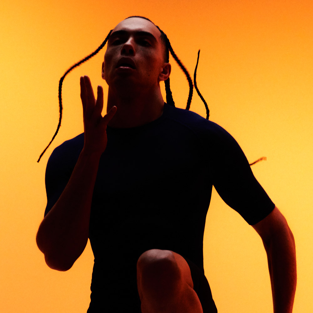 Train with the new compression clothing set for men A man with braids does a knee lift in front of an orange background