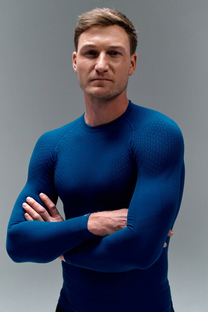 A man stands with crossed arms in a tight-fitting blue athletic top against a grey backdrop. The textured fabric emphasizes muscle definition and radiates strength, confidence, and performance.