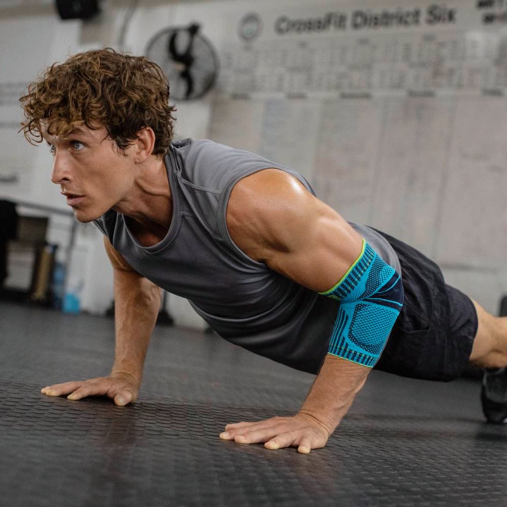 Show your strength with every push-up! A fit man with a muscular upper body performs push-ups in a gym with focus. He is wearing a blue and black elbow support on his right arm for stability and support during his workout.