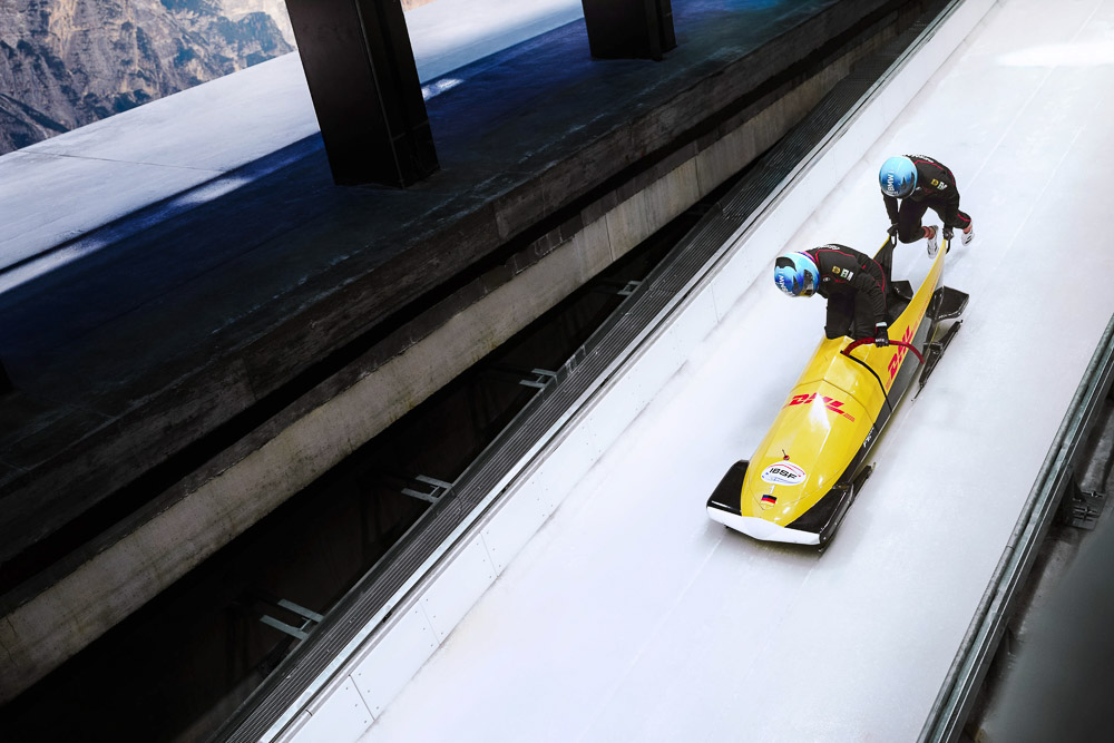 Two female bobsleigh athletes push a yellow two-man bobsleigh along an icy track, captured from an elevated angle. Both athletes wear helmets and racing suits, with the ice channel running between concrete walls in a mountainous setting.