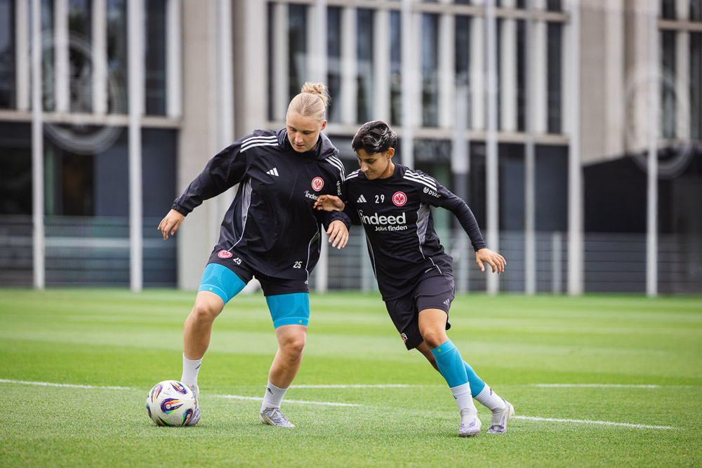 Two Eintracht Frankfurt players training in a duel on the football pitch, both wearing Bauerfeind compression sleeves on their knees.