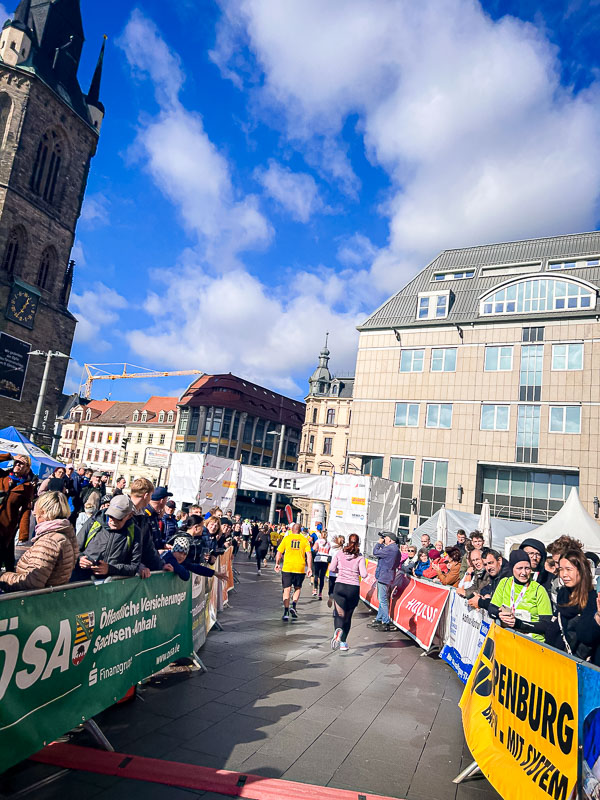 The final meters before the finish line at the half marathon in Halle. Runners are crossing the finish line under blue skies, cheered on by the audience.