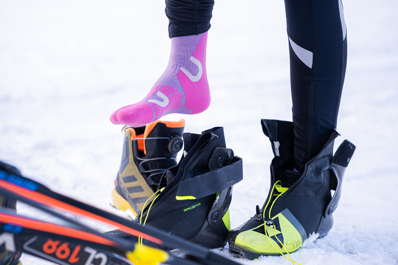 A close-up of cross-country skiing gear: An athlete puts on neon pink functional socks while high-quality skiing boots and skis lie on the snow beside her.
