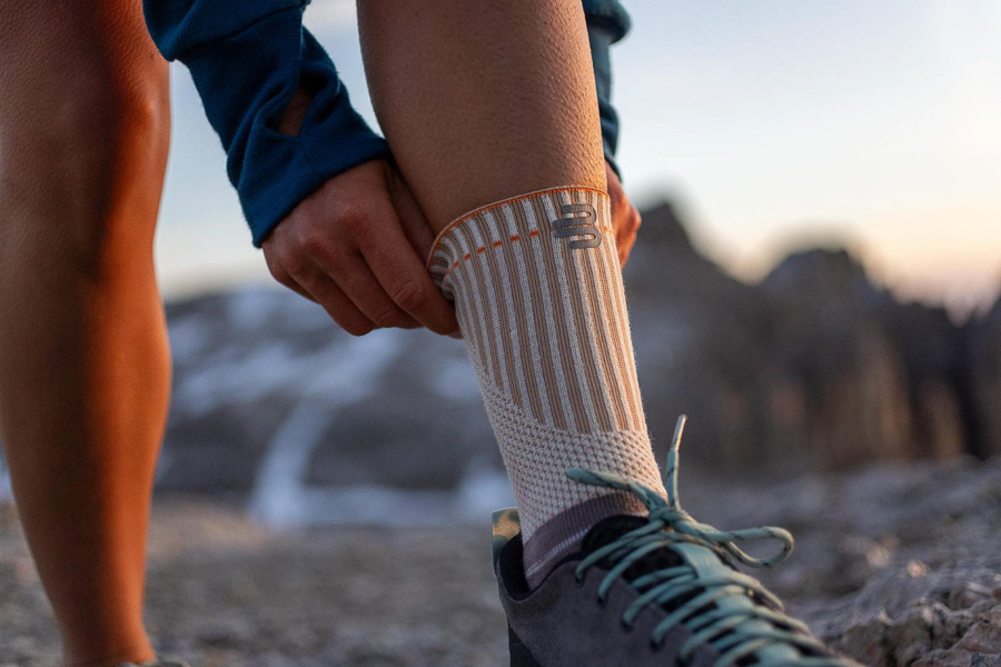 A hiker adjusts their specialized outdoor bandage with reinforced cuff. Mountains and a sunset are visible in the background.