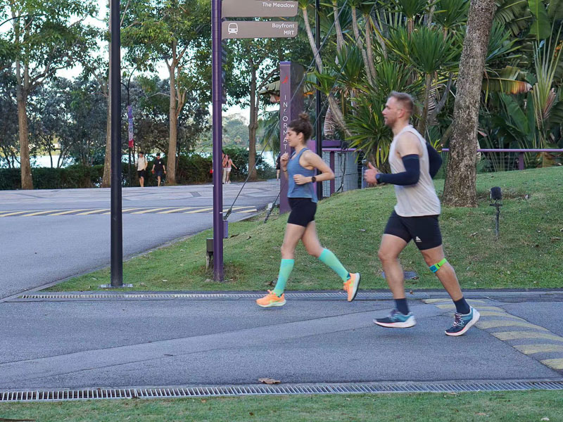 Zusammen stark: Lauftraining für die Fitness Challenge Zwei Personen joggen entlang eines städtischen Parks auf einer asphaltierten Straße. Ein Mann in einer hellen Laufkleidung und einer Kniebandage läuft mit einer jungen Frau, die grüne Kompressionssocken trägt.