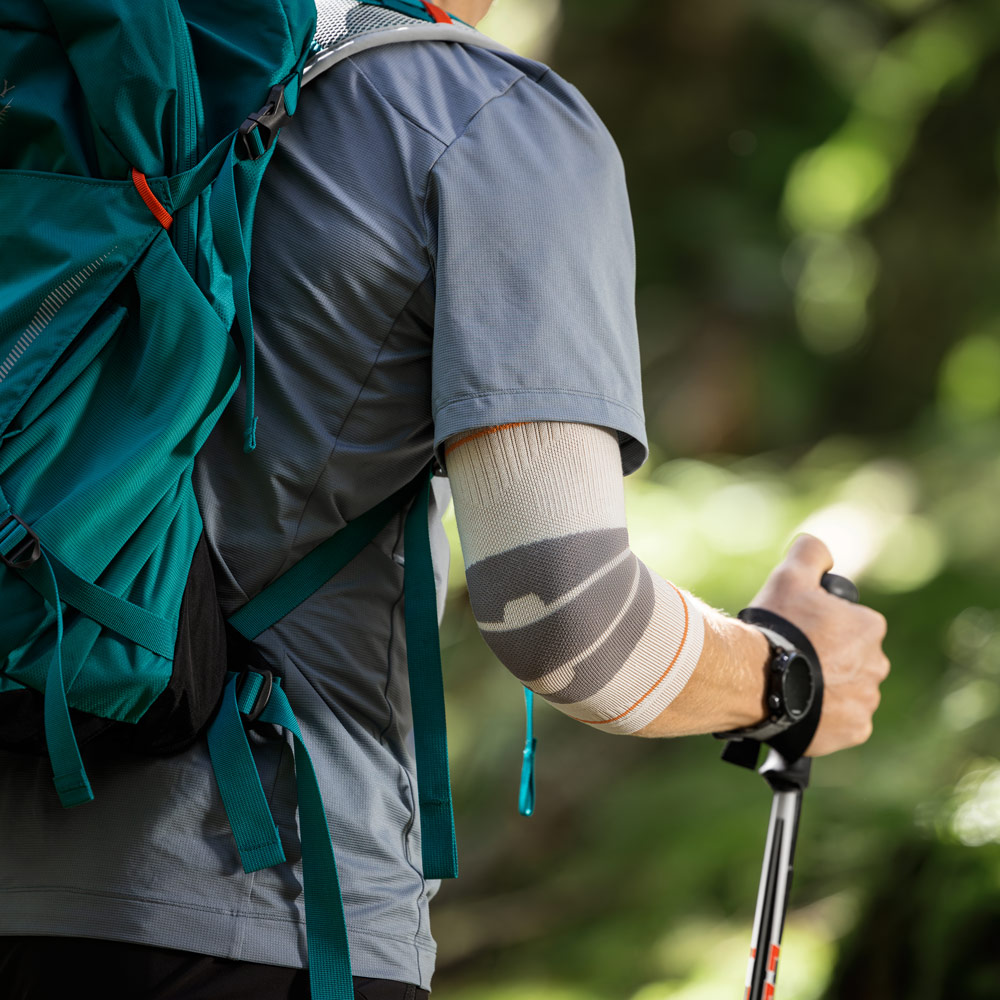 A hiker walks along a narrow forest trail using trekking poles for support. On his right arm, he wears an Outdoor Compression Elbow Support in sandstone tones that closely fits around the elbow.