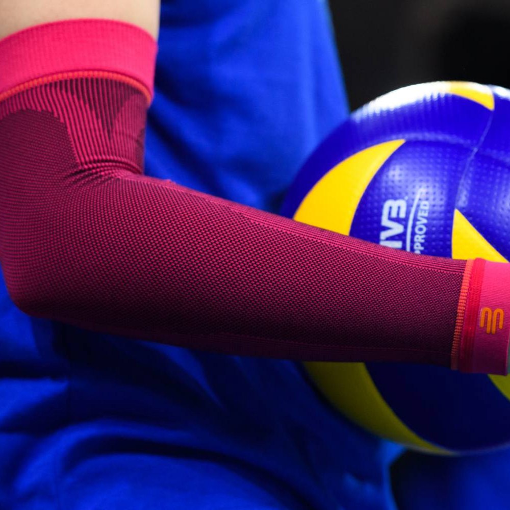 Close-up of a pink volleyball armsleeve on a player’s arm with a yellow and blue volleyball in the background.