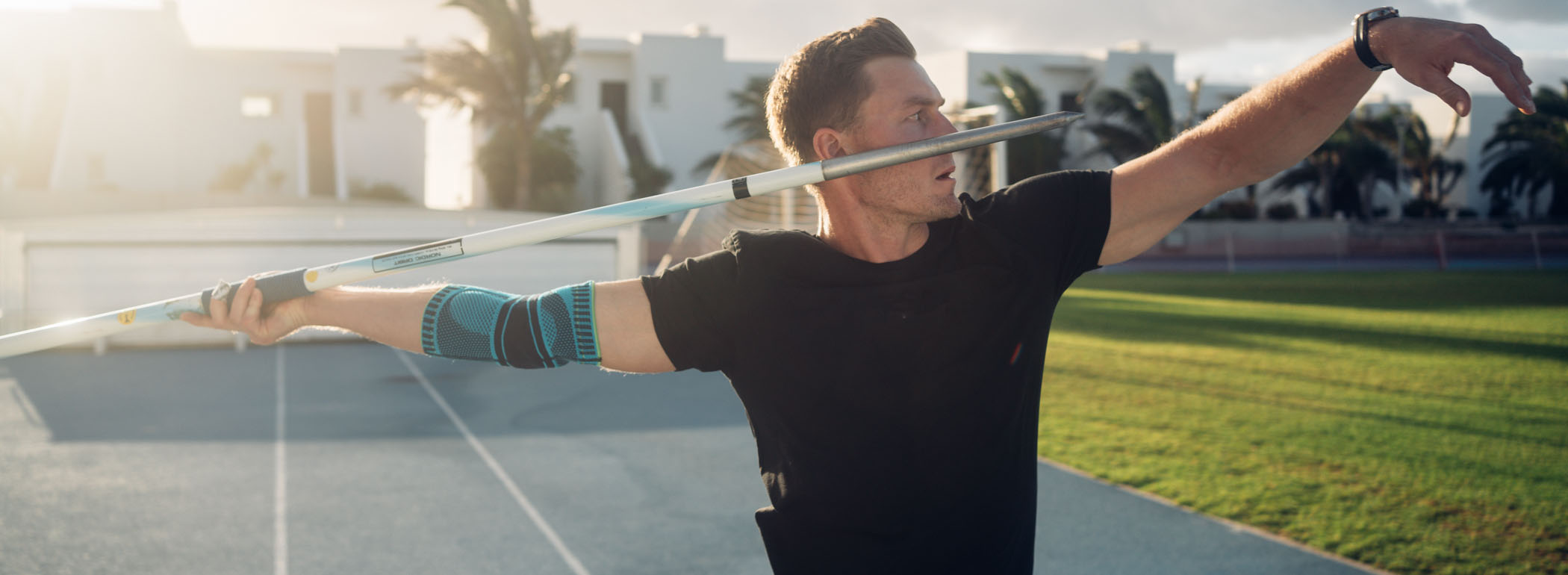 A javelin thrower in the stadium preparing for a powerful throw. He is wearing a blue elbow support brace on his right arm, providing stability, compression, and optimal protection during intense throwing motions.