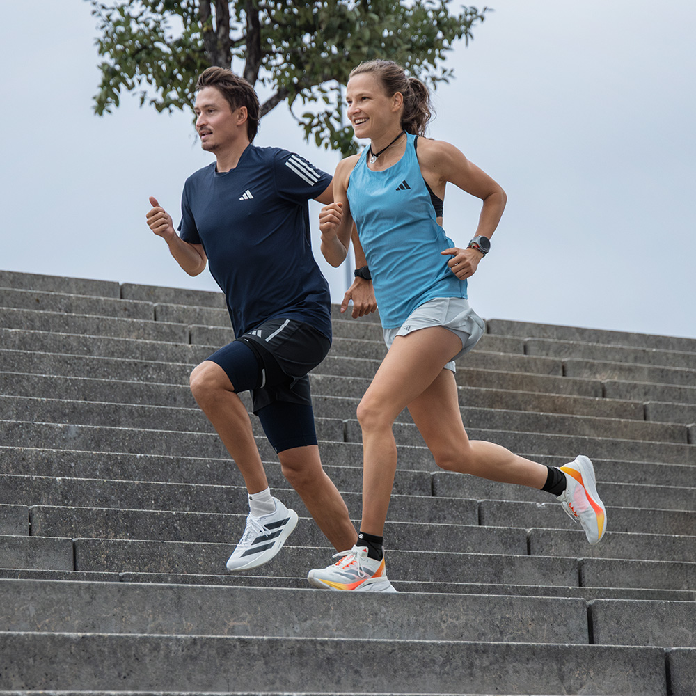 A sporty woman and a man are running down a wide stone staircase together. Both wear adidas sportswear and shoes.