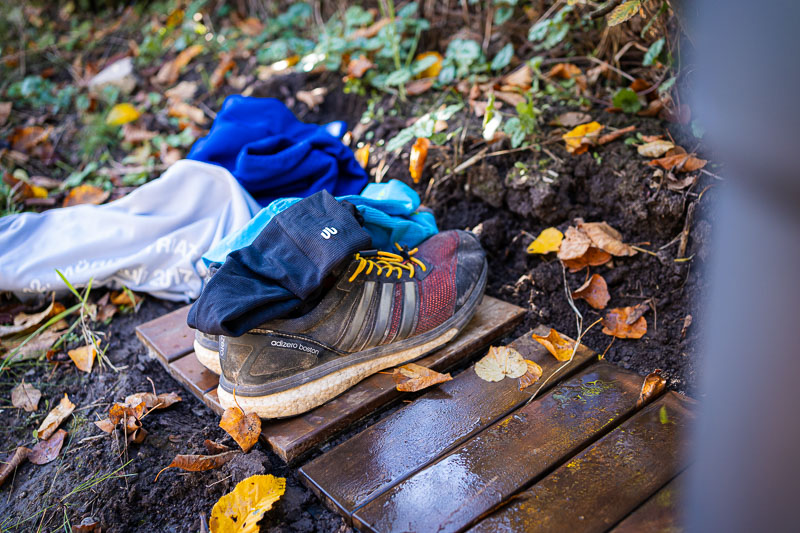 A pair of running shoes with yellow laces on a wooden board. Nearby, a pile of clothes including a blue towel, a grey jacket, and black gloves. Autumn leaves are scattered on the ground.