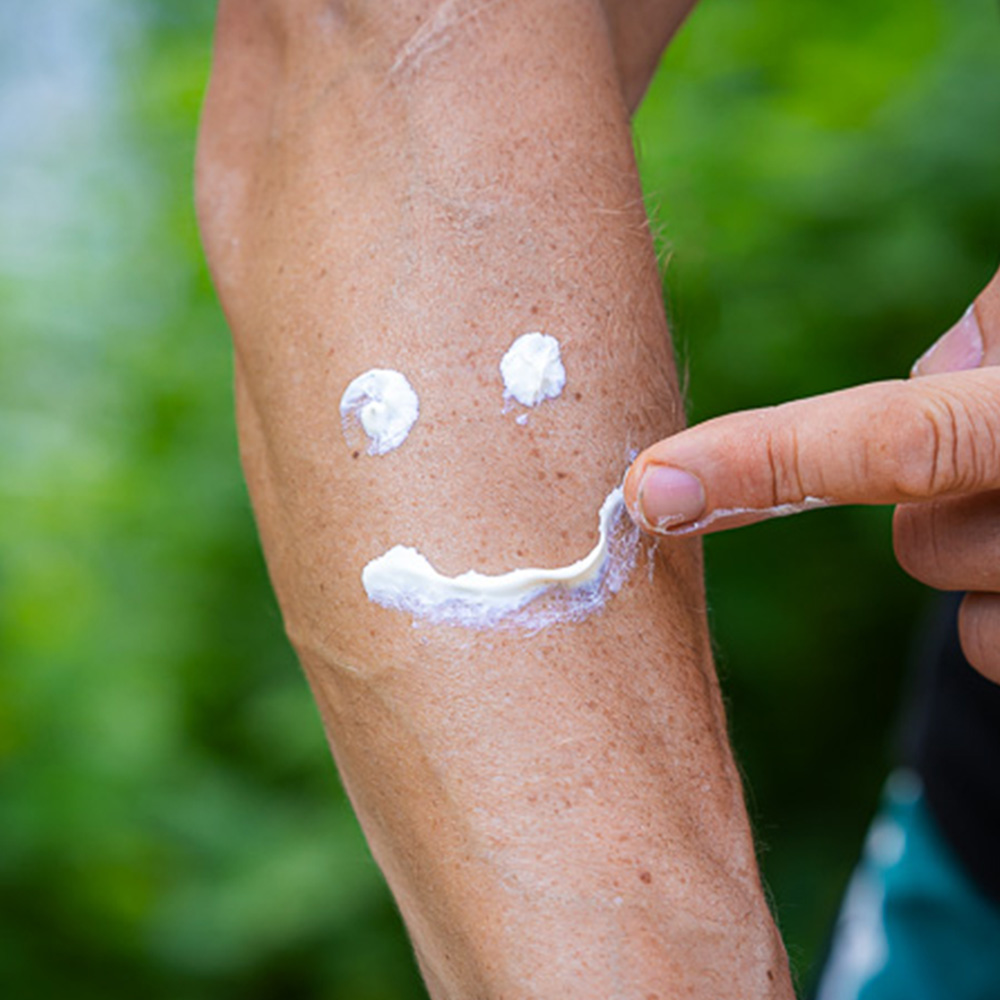 Sunscreen from Bauerfeind Sports is applied on an arm, forming a smiley face. A hand points to the "mouth" of the smiley. The scene takes place outdoors, surrounded by green nature.