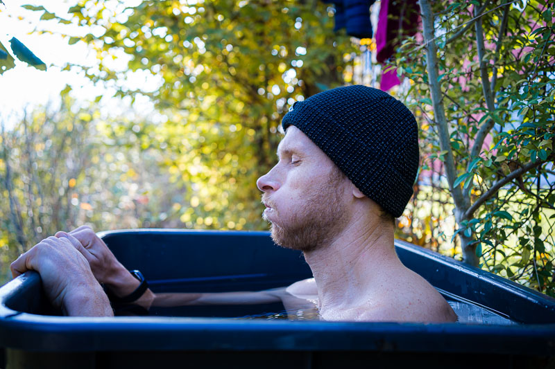 A man wearing a dark knit beanie sits up to his shoulders in an outdoor ice bath. He is surrounded by autumn vegetation and has a focused expression as he breathes deeply, adjusting to the cold stimulus.