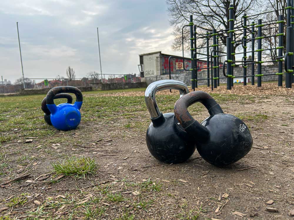 Several kettlebells are placed on bare ground in an outdoor training park, with a calisthenics rig visible in the background. The image shows a functional setup for HYROX training without a gym, where strength and carry exercises can be done outdoors.
