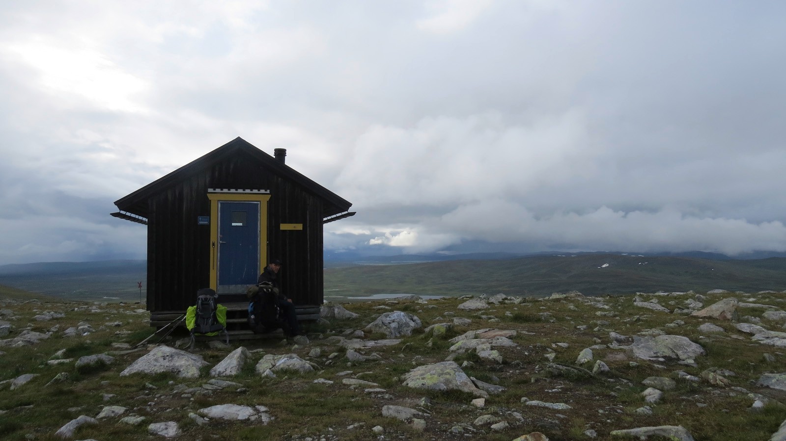 A lonely hut in the mountains in the middle of groving meadows with small rocks in the background the dense cloud cover and a small mountain lake