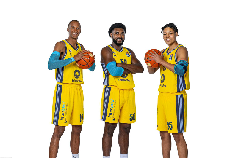 Three Alba Berlin basketball players in their yellow team jerseys posing with basketballs against a neutral white background. Each player smiles while holding a basketball with a relaxed, professional demeanor.