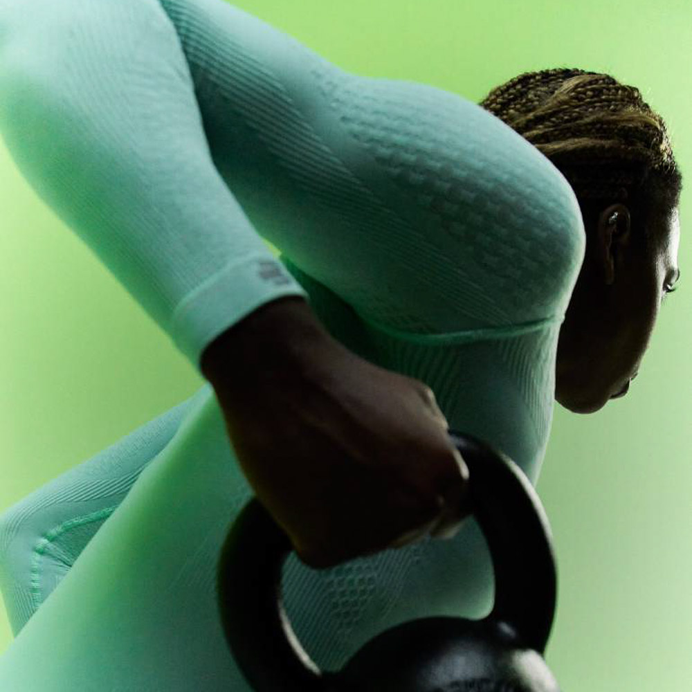 Athlete in a mint green compression top and dark blue compression trousers doing arm training Athlete in mint green compression top and dark blue compression trousers stretches her arm upwards while holding a dumbbell in her hand