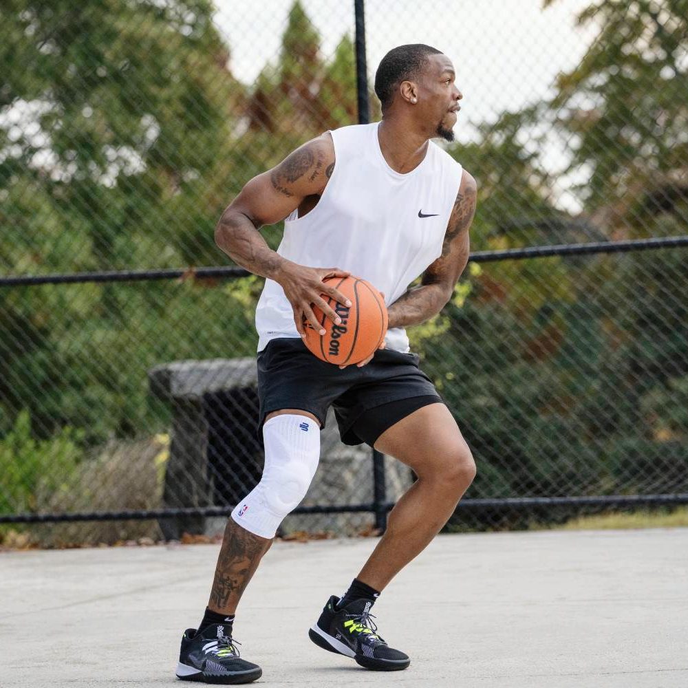 Basketball player in a white jersey preparing to shoot. His right knee is supported by a white compression brace, standing on an outdoor court with hoops in the background.