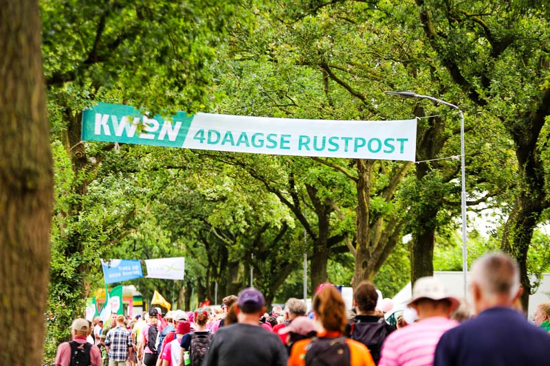 Numerous participants pass a rest stop during the 4Daagse walk. A large banner above them reads "KWBN 4DAAGSE RUSTPOST."