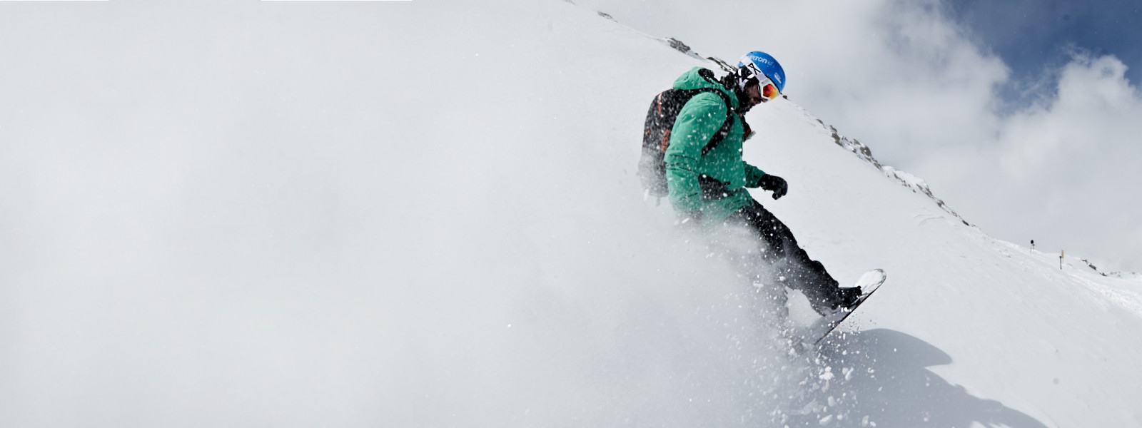 Snowboarders with a green jacket whirls snow on which the entire left side of the picture is dyeing white when braking