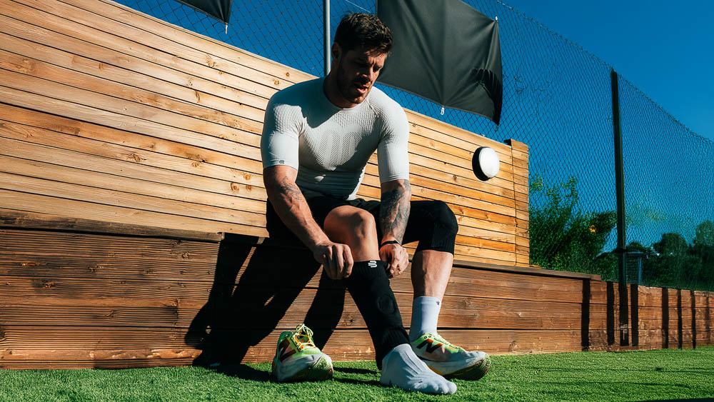 Athlete sits outdoors on a wooden bench and pulls on a black sock or compression layer over his lower leg; he wears a fitted white performance shirt and prepares for training in the sun.