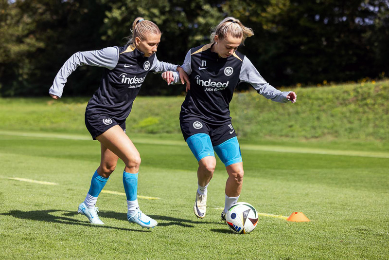 Two Eintracht Frankfurt players battle for the ball during a training match. Both are wearing black training gear with the Eintracht logo and blue knee supports from Bauerfeind Sports.