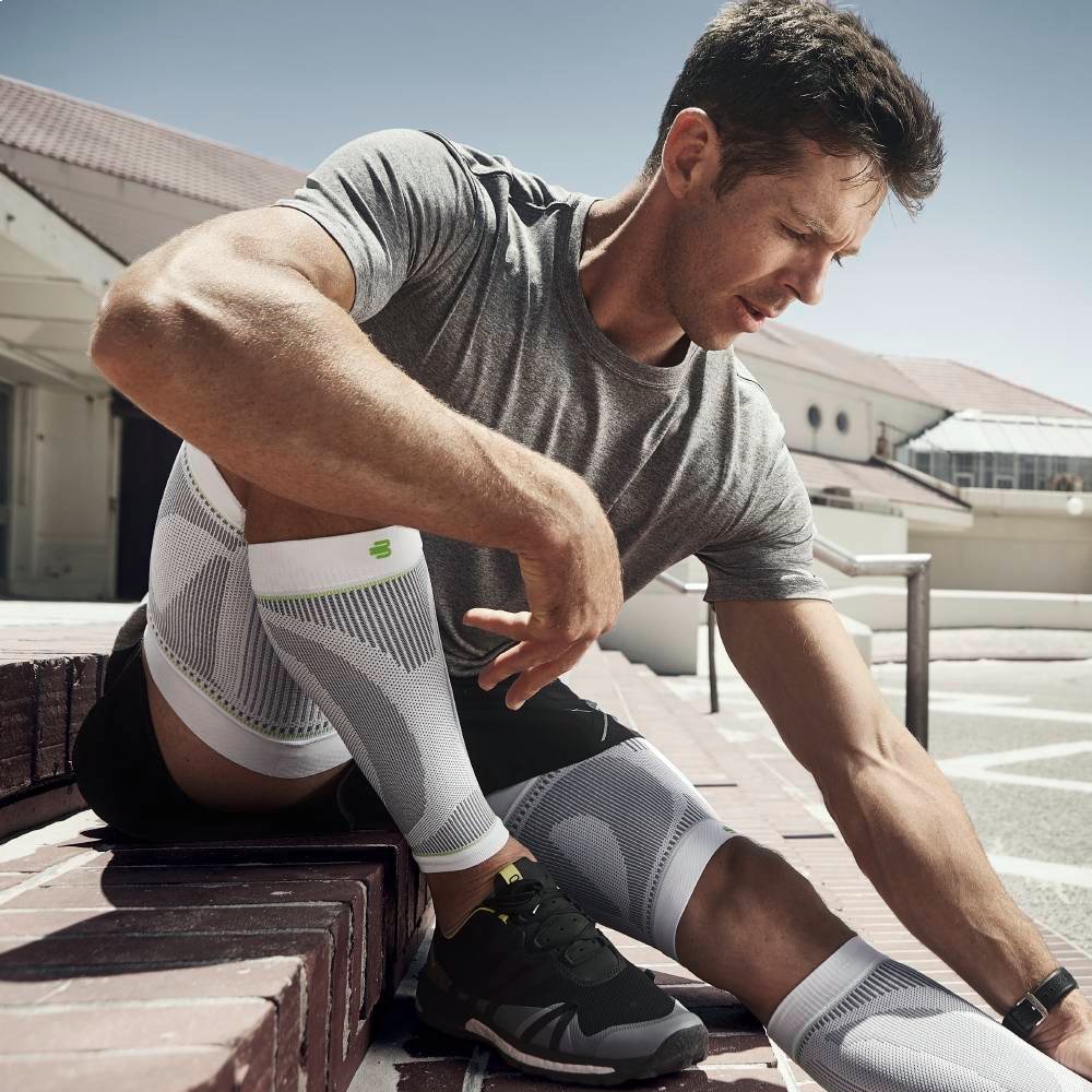 A fit man sits on a staircase outside in the sun, wearing white compression sleeves, seemingly recovering from an intense run. He takes a deep breath, mentally preparing for his next challenge.