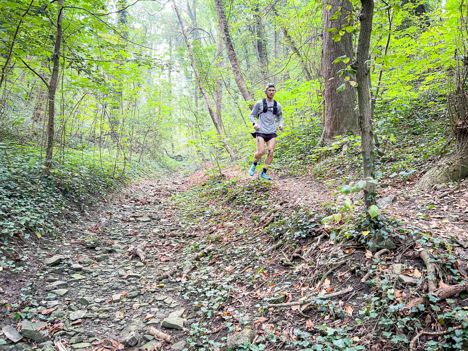 A trail runner running on a rocky path through a dense forest. The surroundings are green and peaceful, perfect for intense outdoor training.
