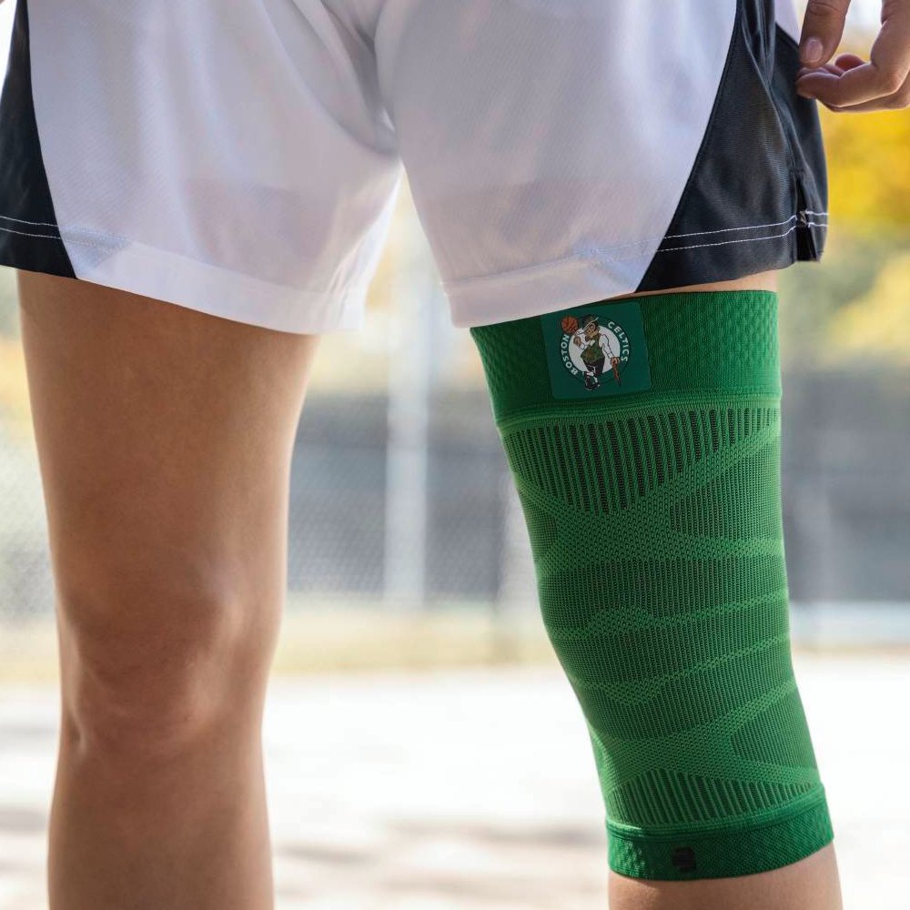 Close-up of a player wearing a green Boston Celtics knee support on a basketball court. The brace features the team logo and fits ergonomically around the knee.