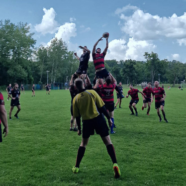 Ein Rugby-Spiel in vollem Gange auf einem grünen Rasenfeld. Ein Spieler in rot-schwarzem Trikot wird von Teamkollegen hochgehoben, um den Ball zu fangen, während der Schiedsrichter in gelber Kleidung die Szene beobachtet.