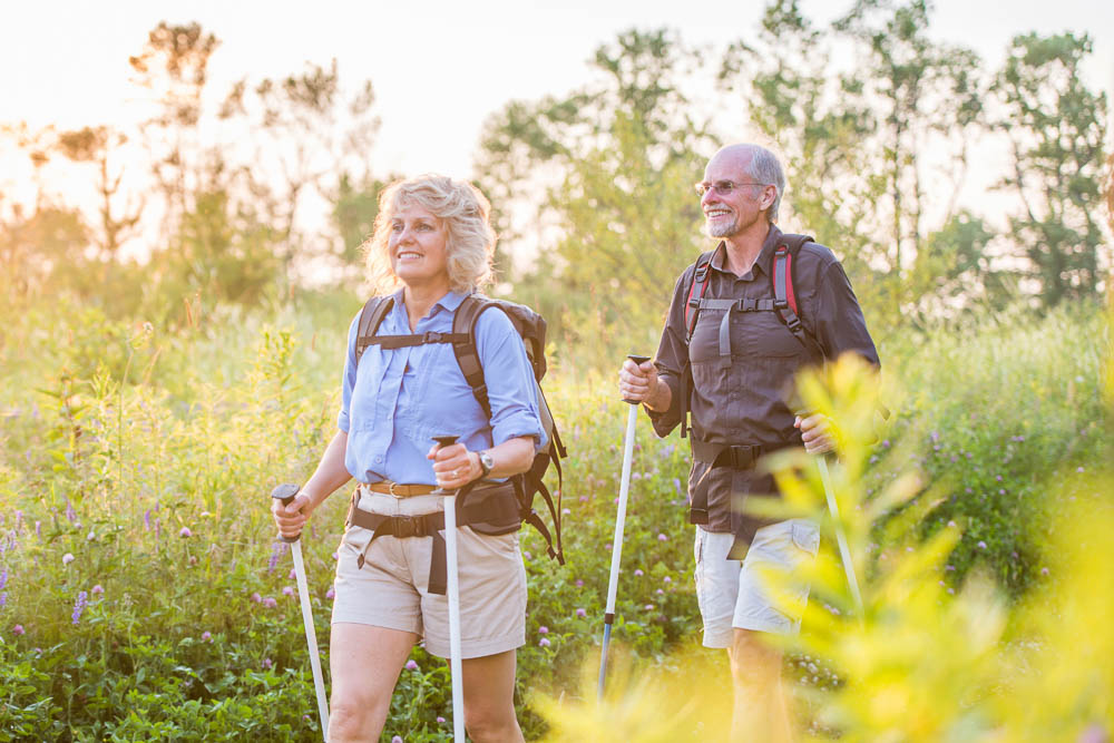 An older couple hikes with trekking poles through a blooming summer meadow in the soft evening light. Both wear lightweight hiking outfits and backpacks, looking content and engaged in nature.