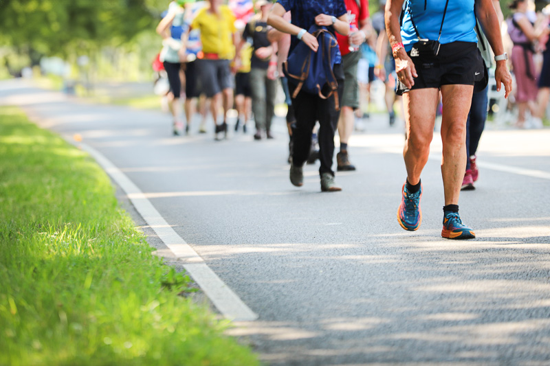 A group of hikers walks along a closed-off road in a green area on a sunny day. In the foreground, a man in blue sportswear and running shoes is visible.