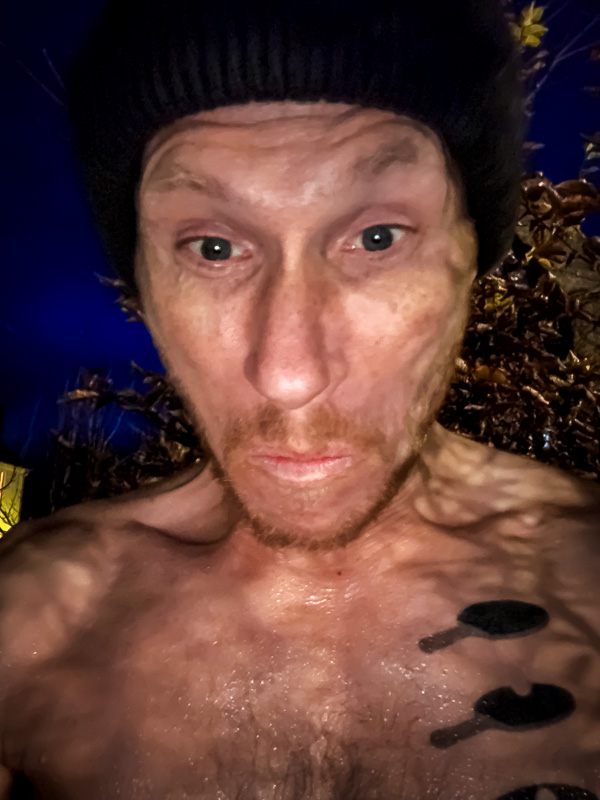 A man with a black beanie and a wet upper body stares tensely into the camera. The background features autumn leaves under a dark evening sky.
