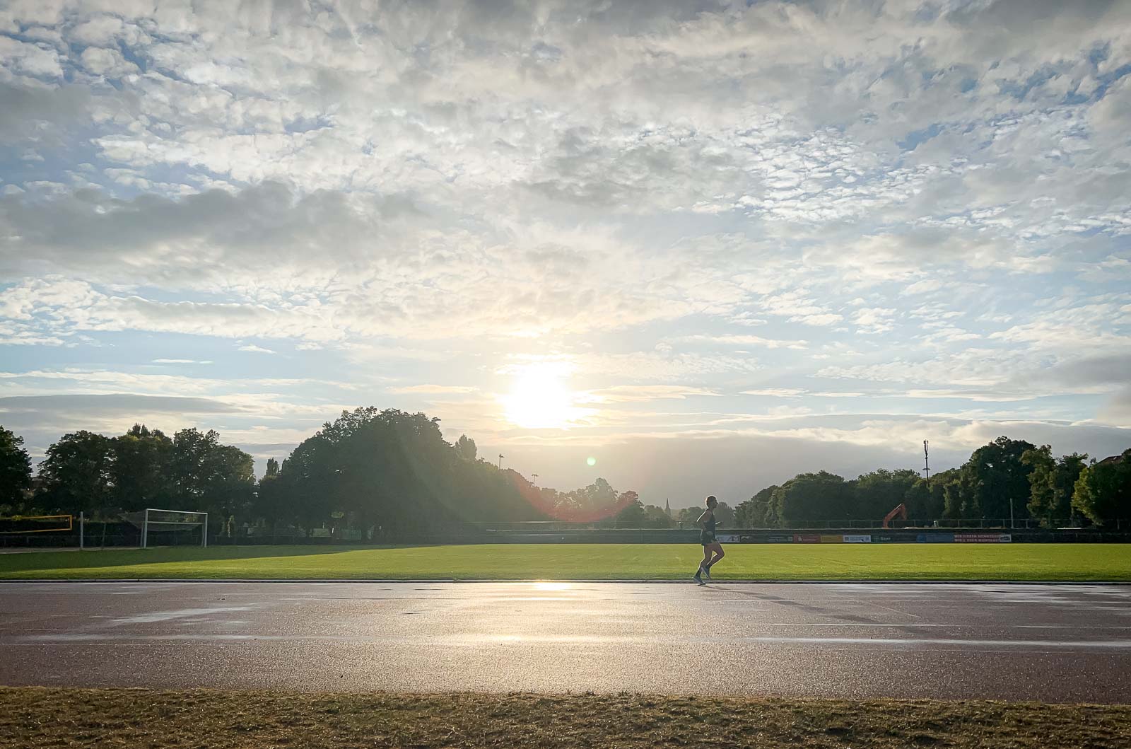 A runner on a wet track at sunrise. The sky is cloudy, but the sun is breaking through as the runner completes his lap.