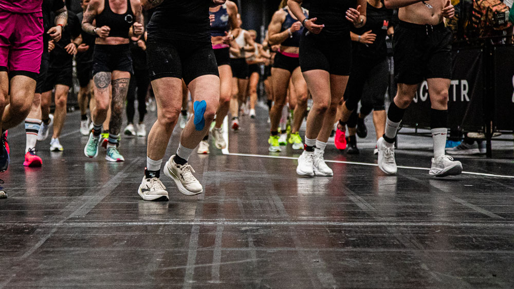Dynamic close-up of the legs of several athletes running on a dark indoor track during a HYROX competition; sports shoes, race gear and blue kinesiology tape on one leg highlight intense movement, endurance, strength and determination.