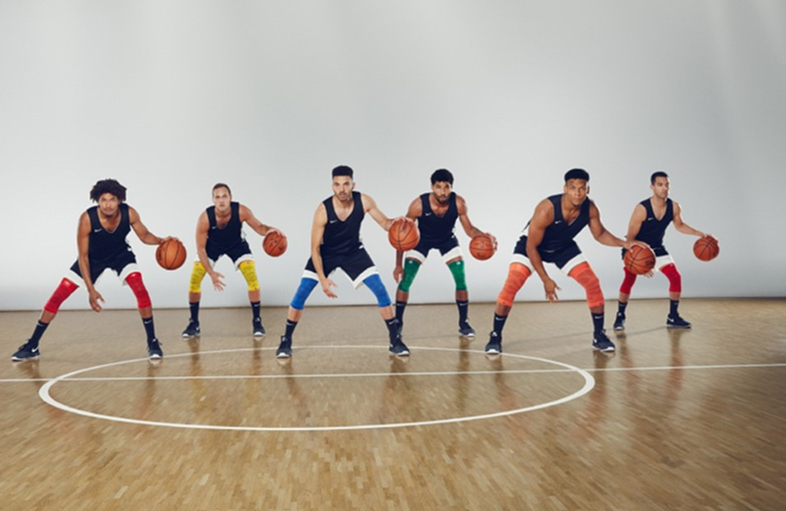 Six basketball players stand in a gym, lined up across the court in a ready stance, each holding a basketball. They wear black jerseys, white shorts, and colorful knee compression sleeves in red, yellow, blue, green, and orange.