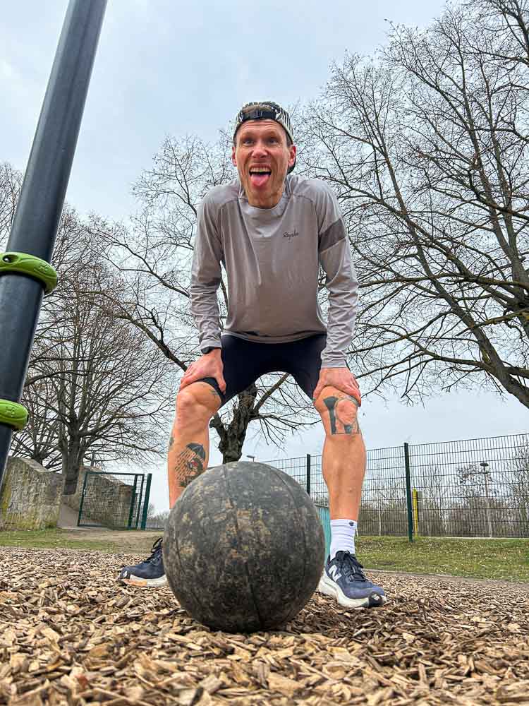 An athlete stands in an outdoor training park with hands on knees above a medicine ball after an intense session. The image captures fatigue after a functional workout typical of HYROX training without a gym.