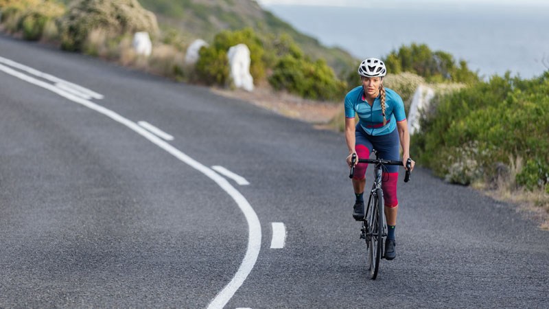 Woman with Armsleeves and knee band in the background of the image runs on a mountain bike over a path between high grass